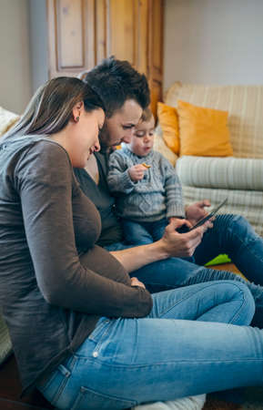 Family with small child and pregnant mother looking at the tablet sitting on the carpet