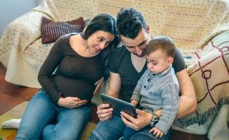 Family with small child and pregnant mother looking at the tablet sitting on the carpet