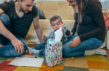 Father and pregnant mother with their little son opening a gift in the living roomの写真素材