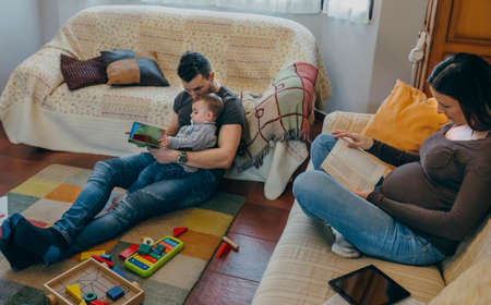 Young father reading a book to his little son while the pregnant mother readsの写真素材