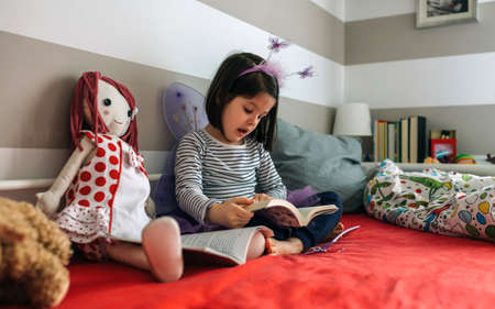 Little girl disguised as a butterfly sitting on the bed reading a book to her rag doll