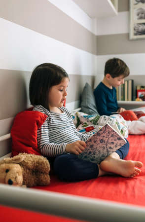 Girl and boy reading a book each sitting on the bed. Selective focus on girl in foreground