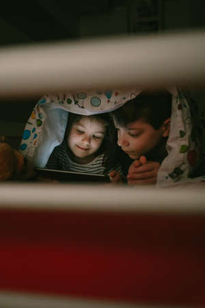 Brother and sister lying on the bed looking at the tablet in the dark