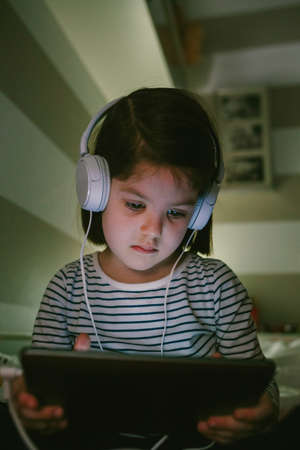 Little girl with headphones looking at the tablet sitting on the bed at nightの写真素材