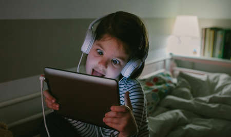 Funny girl grimacing with headphones looking at the tablet sitting on the bed at night