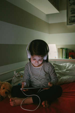 Little girl with headphones looking at the tablet sitting on the bed at night