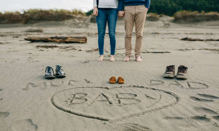 Mum, dad and baby written on the sand of the beach next to their shoes with the legs of the parents behindの写真素材