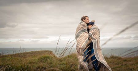 Portrait of young beautiful couple embracing under blanket in a cold day with sea and dark cloudy...