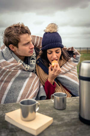 Portrait of young beautiful couple under blanket eating muffin in a cold day with sea and dark...