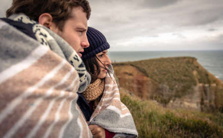 Closeup of young beautiful couple under blanket in a cold day looking the sea with the dark cloudy sky on the backgroundの写真素材
