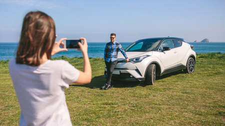Young woman taking a picture of guy leaning on a car near the coast