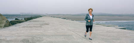 Senior sportswoman with headphones running by sea pierの写真素材