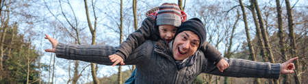 Happy man giving piggyback ride to smiling kid and raising their arms into the forest