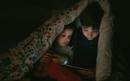 Brother and sister sitting on the bed looking at the tablet in the darkの写真素材