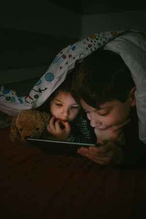 Brother and sister lying on the bed looking at the tablet in the darkの写真素材