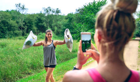 Girl taking a picture of a friend with trash bags after plogging. Selective focus on girl in...