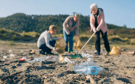 Group of senior volunteers picking up trash on the beach. Selective focus on plastic bottle in...