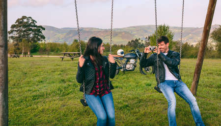 Young man taking a picture of his girlfriend sitting on the swings in a recreational areaの写真素材