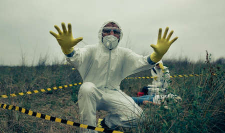 Man with bacteriological protective suit prohibiting entering cordoned off area while his partner looks at a corpseの写真素材