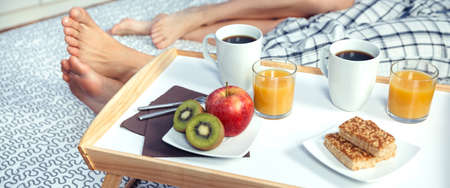 Closeup of healthy breakfast served on a wooden tray ready to eat and couple legs over a bed in the background. Healthy food and home lifestyle concept.の写真素材