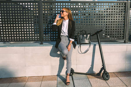 Businesswoman talking on the mobile sitting on a wall with her scooter nextの写真素材