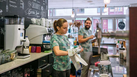 Young waitress with mask cleaning glasses in a coffee shop next to her coworker
