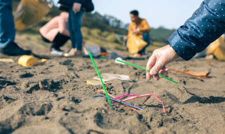 Woman hand picking up straws on the beach with group of volunteers working in the background....