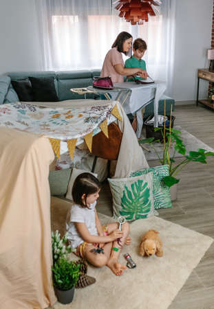 Mother teaching her teenage son to iron while her daughter play camping at home in the living roomの写真素材