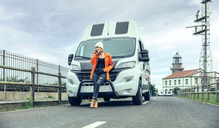 Young woman leaning on camper van on the road calling by mobile for a breakdown with lighthouse in...
