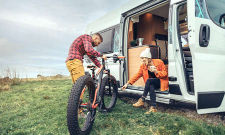 Woman drinking coffee sitting at the door of a camper van and man checking fat bikeの写真素材