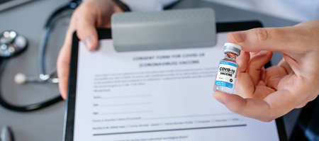 Detail of female doctors hand showing coronavirus vaccine vial and consent form. Selective focus on...
