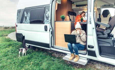 Woman teleworking drinking coffee sitting in the door of a camper van while her partner works insideの写真素材