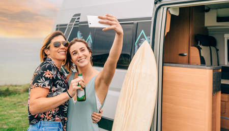 Two happy young women taking a selfie with their cell phone next to their camper van during a tripの写真素材