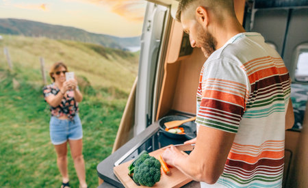 Young man cooking vegetables in a camper van while his wife takes a picture of him with her cell phone. Selective focus on man in foregroundの写真素材