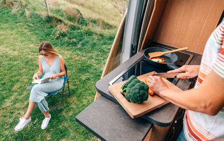 Young man cooking vegetables in a camper van while young woman reads outdoorsの写真素材