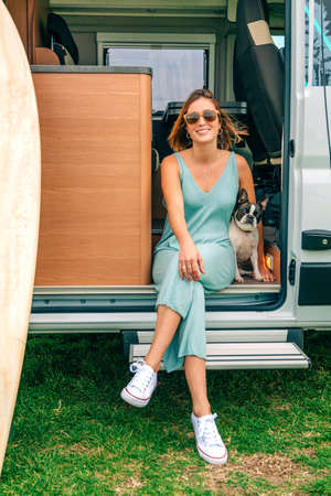 Smiling young woman sitting with her boston terrier dog at the door of her camper van during a tripの写真素材