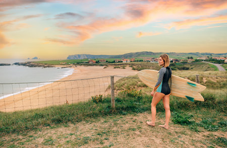 Young surfer woman with wetsuit and surfboard looking at the beach from the shore
