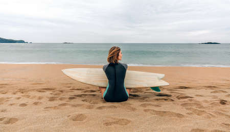 Young surfer woman with wetsuit and surfboard sitting on the sand looking aside on the beach
