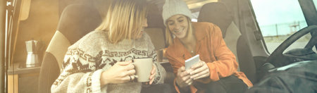 Two women laughing looking mobile with a cup of coffee sitting in the front seat of a camper van