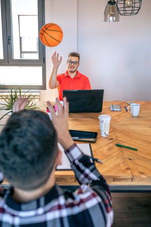 Smiling young businessman searching new ideas in brainstorm while having fun throwing a basket ball to colleague in a coworking office. Relax and creativity time at office.の写真素材