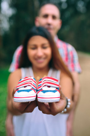 Smiling latin pregnant woman showing small baby sneakers while her partner holding her belly on background. Selective focus on sneakers in foreground.の写真素材