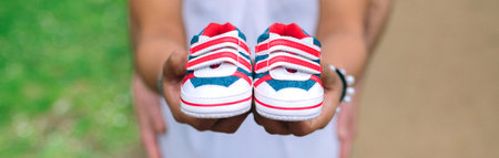 Unrecognizable pregnant woman showing small baby sneakers while her partner holding her belly on background. Selective focus on sneakers in foreground.の写真素材
