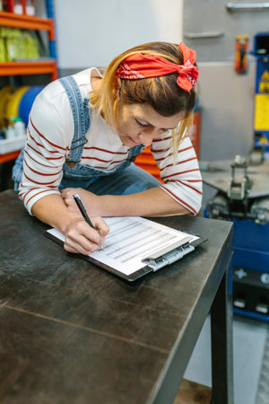 Concentrated woman leaning on workshop table checking stock products of mechanic garage