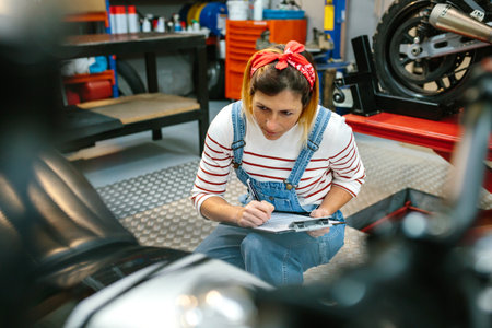 Concentrated mechanic woman with clipboard reviewing damages in motorbike on garageの写真素材