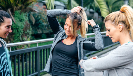 Smiling blonde woman runner tying her hair in a ponytail next to female friends before trainingの写真素材