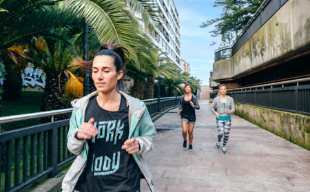 Young brunette woman with ponytail and piercings running ahead of female friends team over a urban runway with palm treesの写真素材