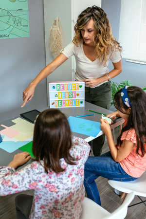 Female teacher explaining to students in classroom how it is a ecologic process while holding lightbox with text reduce, reuse, recycle. Sustainable lifestyle education concept.の写真素材