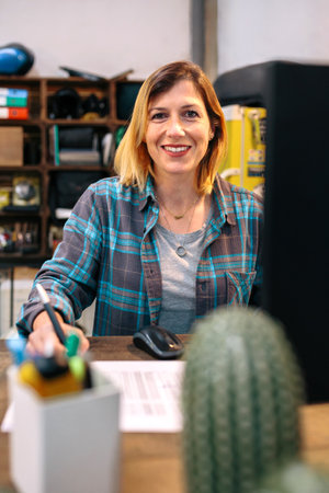 Portrait of smiling blonde young woman filling order forms for ecommerce shop. Cheerful woman with freckles wearing a plaid shirt and working behind of computer monitor in store selling on line.の写真素材