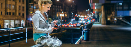 Smiling woman looking her coach messages on mobile app while open bottle of water after training. Banner of female young runner with earphones sitting over banister at night on town. Right copy space.の写真素材