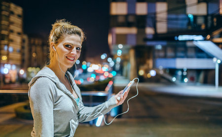 Portrait of smiling young blonde sportswoman listening music on mobile phone application. Female runner with earphones looking at camera after training at night on empty town. Right copy space.の写真素材
